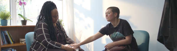 A still from a film scene. Seated woman with cropped hair to the right presses a pad of paper into the lap of a seated woman with gathered braided hair to the left.